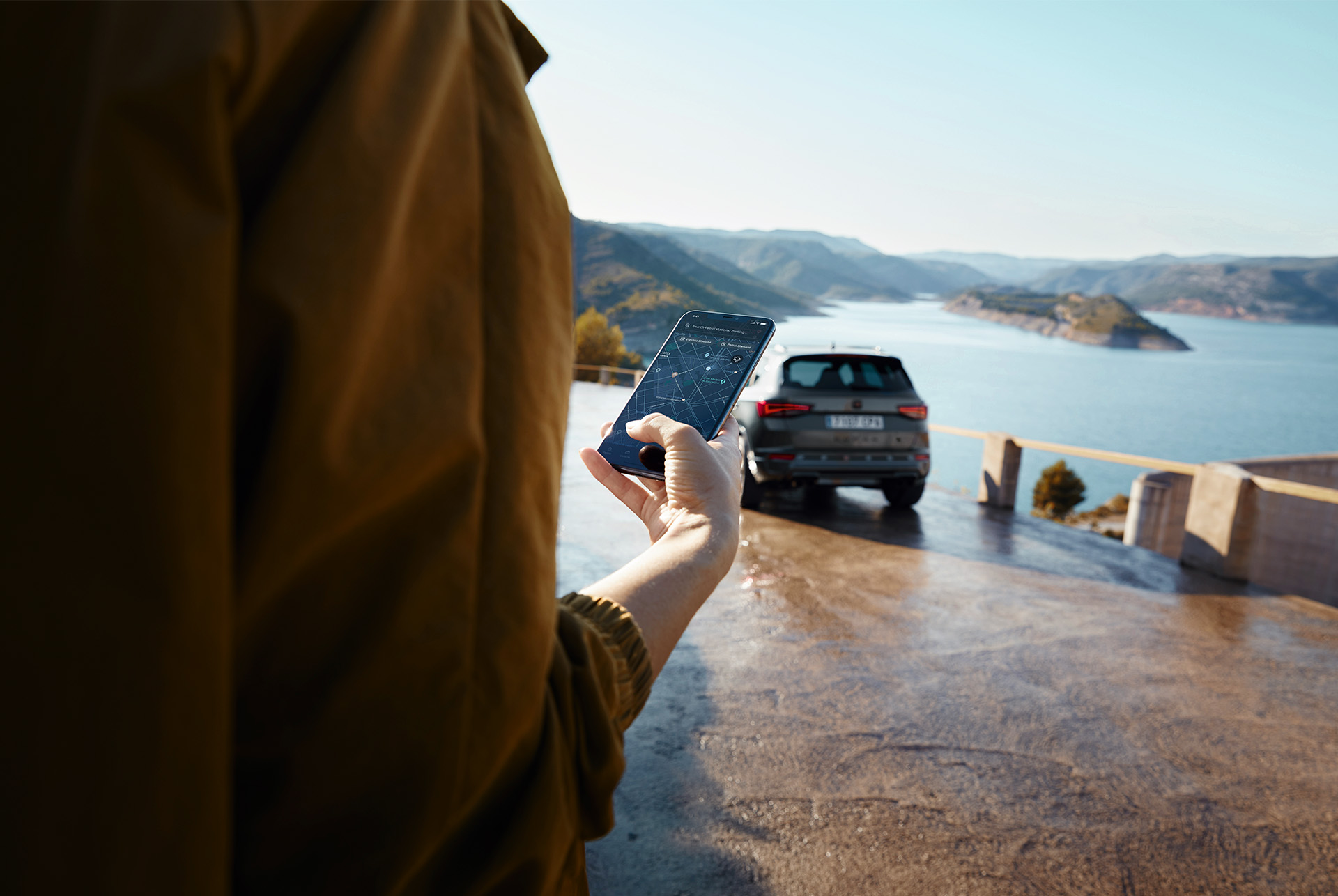 Una persona sostiene un smartphone con el CUPRA Ateca de fondo, aparcado cerca de una vista panorámica de montañas y agua, simbolizando un mundo conectado.