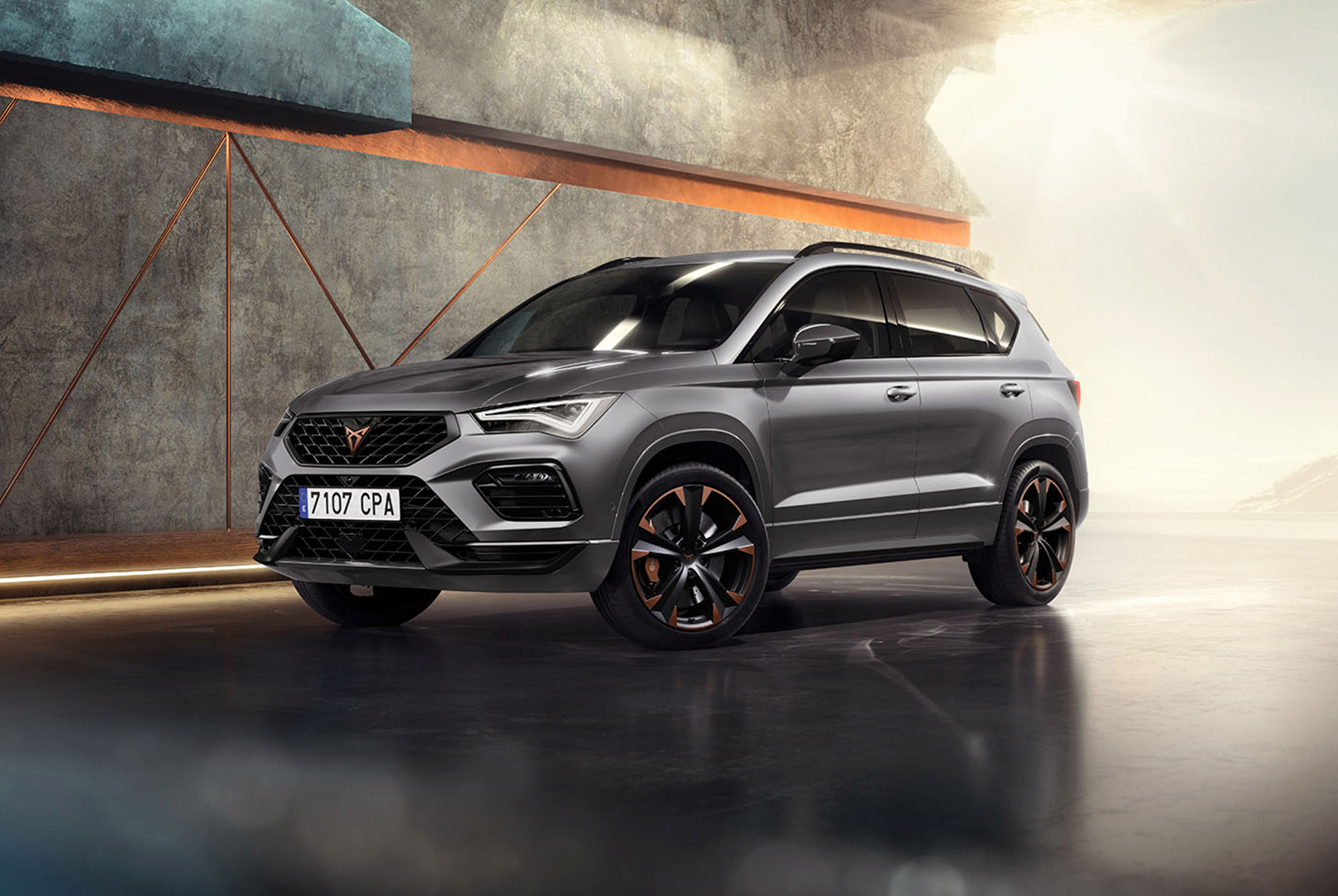 CUPRA Ateca 2024 en gris graphite, un SUV deportivo y compacto aparcado en un entorno moderno e industrial. El diseño del coche presenta líneas aerodinámicas, faros LED y llantas de aleación distintivas con detalles en cobre. En el fondo hay una pared con un diseño geométrico, que contrasta con el elegante diseño del vehículo.